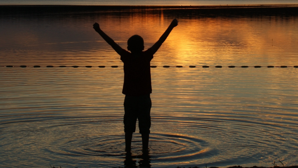 Picture of a boy standing at the edge of a lake at sunset hands in the air celebrating